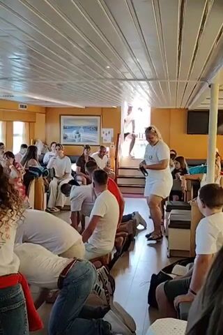 A group of people are gathered in a room with a white, paneled ceiling. Some individuals are kneeling or bent over on the floor, while others are standing. One person, a woman with blonde hair, is standing and appears to be speaking or giving instructions. The room has wooden floors and appears to be on a boat or ship, with windows visible in the background. The people are dressed casually, many in white t-shirts and shorts. There is a painting on the wall depicting a white building with blue accents.