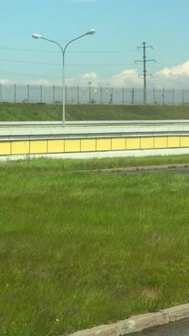 The video shows a view from inside a moving vehicle, looking out at a roadside scene. There is a grassy area in the foreground with a curb visible at the bottom. Behind the grass is a road or overpass structure with yellow barriers. Beyond that is a fence, some trees, and utility poles against a blue sky with some clouds. A road sign is visible on the right side of the frame, indicating a restricted area for towing services. The vehicle is moving, as the background elements shift in perspective.