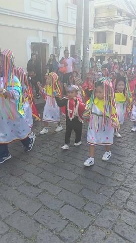 A vibrant parade featuring young children dressed in colorful traditional or festive attire is captured on a cobbled street. The children, appearing to be of preschool or early elementary age, are marching or dancing in a procession.

Many children are wearing white tunics or dresses with yellow tops and elaborate, multi-colored ribbon headpieces. These ribbons, in shades of red, yellow, green, blue, purple, and orange, are attached to their heads and cascade down around their faces, creating a lively and festive appearance. The white garments are adorned with colorful patterns or embroidery near the hem.

Interspersed among the children in white and yellow, several others are dressed in red capes or full red outfits, often with black accents. One child in a red cape and black attire wears a dark, possibly devil-like or mythological creature mask. Another child in a similar red and black ensemble sports a distinctive silver, crown-like headpiece. All the children are wearing white athletic shoes.

The children are moving forward in a somewhat informal line, swaying and stepping to the upbeat music audible in the background. Their expressions range from focused to joyful, indicating active participation in the event.

The setting is an urban street paved with cobblestones. Traditional-looking buildings with light-colored facades line both sides of the street. In the background, numerous adults are visible, some observing the parade, others walking alongside the children. A partially visible sign for a business, displaying the word "REGALOS" (gifts), can be seen on one of the buildings. The scene appears to take place during the day, with bright, ample lighting. The overall atmosphere is festive and celebratory.