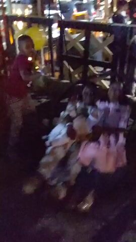 The video shows children playing in an outdoor area at night. There is a yellow balloon visible. The area seems to be a playground or a party venue, evidenced by the presence of people, fencing, and music in the background. Some children are seated on what appears to be a swing-like structure fashioned from tires and a blanket. Another child stands nearby. The scene is darkly lit, and some details are not sharp. There are cars and buildings visible in the background behind the play area.