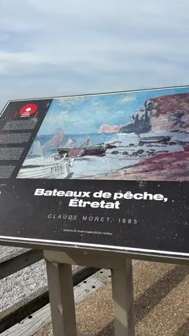The video begins with a close-up of a framed informational sign featuring a painting titled "Bateaux de pêche, Etretat" by Claude Monet, painted in 1885. The sign is outdoors, with a wooden railing visible in the foreground. The camera then pans to the right, revealing a wide view of a pebble beach with numerous people relaxing and walking. Majestic white cliffs, characteristic of Etretat, France, dominate the background. The sea is calm, and the sky is overcast with scattered clouds. Several people are seated on a wooden boardwalk or railing, observing the beach and the cliffs.