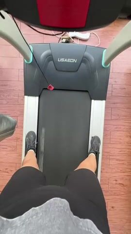 A first-person perspective video shows someone running on a treadmill. The camera is positioned to capture the runner's legs and the treadmill's console. The runner is wearing black shorts and grey athletic shoes. The treadmill is dark gray and silver with a black running belt. The console has various buttons and a digital display. In the background, a window offers a view of a cityscape with tall buildings. The video transitions from a close-up of the runner's legs to a view of the treadmill console and the cityscape beyond. The video appears to be shot indoors, possibly in a gym or apartment with a view.
