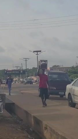 A medium shot shows a man walking to the right along a curb while balancing a box of fruit on his head. The man is wearing a red t-shirt and black shorts, and he's barefoot.  The street is busy with cars, including a maroon van, a silver sedan, and several others in the background. A woman walks on the left side of the road, and various street lights and power lines are visible overhead. The sky is overcast.