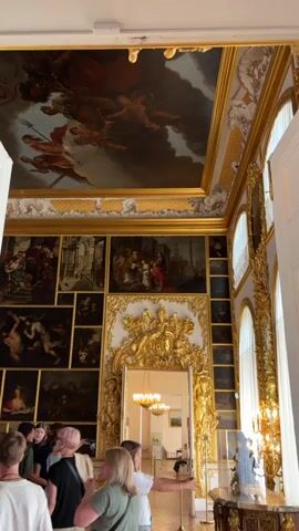 The video shows a group of people, mostly young adults, looking at artwork in a richly decorated room. The room has a high ceiling adorned with a large fresco and walls covered in numerous framed paintings. A large, ornate golden frame surrounds a doorway or mirror in the center of one wall. The paintings on the wall are arranged in a grid-like pattern. One person in the foreground is pointing towards the artwork. The room is well-lit, with natural light streaming in from large windows. The overall impression is of a museum or a historical palace.