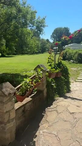 The video shows a sunny day in a park. The camera pans across a green lawn, trees, a stone wall with flower boxes, and a paved path with a wooden fence. A small stream runs alongside the path. In the distance, there are some people walking on the path. The overall impression is one of a peaceful and scenic outdoor environment.