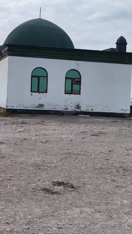 A white building with a dark green dome is seen from the side. The building has two arched windows with green glass. The ground in front of the building is gravelly. The sky is cloudy.