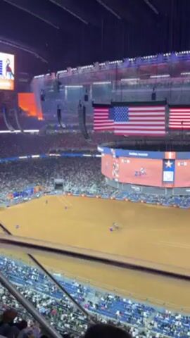 The video shows a crowded arena with a large dirt arena in the center. There are people seated in the stands surrounding the arena. Two large American flags hang from the ceiling. Above the arena, a large video screen displays images and information. A man in a pink shirt stands in the foreground, partially obscuring the view. The arena is identified as NRG Stadium on a sign. The video appears to be taken from a high vantage point within the arena. An event, possibly a rodeo or equestrian competition, is taking place in the arena.