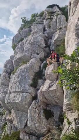 The video begins with a medium shot of a person sitting on a rocky ledge, wearing a helmet and a backpack. The camera then pans to the left, revealing a vast, mountainous landscape with jagged rocks and scattered trees. The sky is partly cloudy, with the sun casting rays of light through the clouds. The camera continues to pan, showing more of the rugged terrain and distant mountain ranges. Finally, the video focuses on a small tree with red leaves in the foreground, with the mountains and sky in the background.