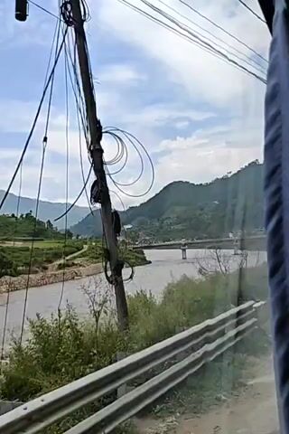 A scenic view of a wide, muddy river flowing through a valley. The river has a strong current with visible ripples and small waves. On the left bank, there are terraced fields and some vegetation. Across the river, a bridge spans the water, with vehicles visible on it, suggesting it's a road bridge. On the right bank, a steep, green hillside rises, dotted with some buildings and trees. The sky above is partly cloudy with a bright, hazy sun. The perspective suggests the video is being filmed from a moving vehicle, likely a bus or car, as indicated by the slight blur and the edge of a window visible in the foreground. The overall environment is mountainous and rural.