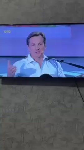 The video shows a man on a television screen. He is wearing a white shirt and blue pants and playing a black guitar with the name "ELVIS" written on it in white. The man is gesturing with his right hand and looking at the camera. Behind him is a stage with blue and white lighting. The TV is mounted on a wall with a gray and white patterned wallpaper. A few hands are visible at the bottom of the screen, suggesting someone is reaching up to interact with the TV. The CTC logo is visible in the top left corner of the screen.