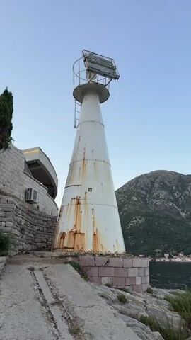 A white conical lighthouse with visible rust stains stands on a rocky shore. The lighthouse has a metal ladder attached to its side and a light fixture at the top. A stone wall and a building with an air conditioning unit are visible to the left of the lighthouse. Mountains are in the background, and a body of water is to the right. The sky is clear and blue.