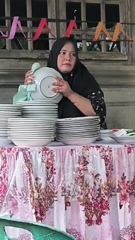 A woman wearing a black hijab and a patterned dress is stacking white plates on a table covered with a floral tablecloth. She is holding one plate and appears to be inspecting it. Another woman, also wearing a hijab and a long dress, stands to the right of the table, facing the first woman. The background features a rustic building with a decorative window and colorful streamers hanging above. Several green plastic chairs are visible in the foreground.