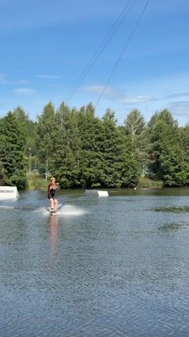 A person wakeboarding on a lake is pulled by a cable. The person is wearing a yellow helmet and an orange life vest over a dark shirt and black shorts. The wakeboarder is creating a spray of water behind them. In the background, there are trees and a clear blue sky with some clouds. There are also some white rectangular structures in the water.
