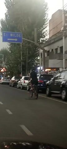 The video shows a person riding a bicycle on a city street. The person is wearing dark clothing and white sneakers. The bicycle has red rims. The street is lined with parked cars. A sign points to "Est. Wilde". There are trees and buildings in the background. The video is shot from behind the cyclist, likely from a moving vehicle. There is background music.