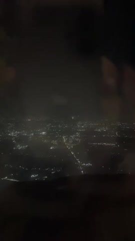 The video, captured from an airplane window at night, shows a cityscape illuminated by numerous lights. The lights appear to be streetlights and building lights spread across a wide area. Clouds partially obscure the view, creating a hazy effect. The lower portion of the frame is dark, suggesting the edge of the airplane's wing or the fuselage.