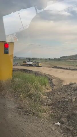 A video taken from inside a vehicle shows a construction site outside. A yellow structure is on the left with a red traffic light. In the background, there are two pieces of construction equipment, a white truck and a yellow excavator or loader. The ground is dirt and there is grass and some foliage. The sky is cloudy and the overall scene appears to be outdoors during the day. There is a reflection on the window that obscures parts of the scene.