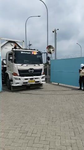 A white tank truck is parked in front of a blue gate. A security guard wearing a yellow vest and a white helmet stands near the gate, looking at their phone. The truck has the words "Dehengon PDI" written on the windshield and "MARUNA JAYA" on the side. The ground is paved with cobblestones. The sky is cloudy.