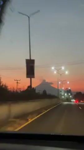 The video shows a view from a moving vehicle on a highway at dusk or dawn. The sky is a gradient of orange and gray. Streetlights illuminate the road, and power lines run overhead. In the distance, a mountain peak is visible against the colorful sky. Other vehicles are visible on the road, including cars with red taillights. The camera appears to be inside the vehicle, looking out through the windshield.