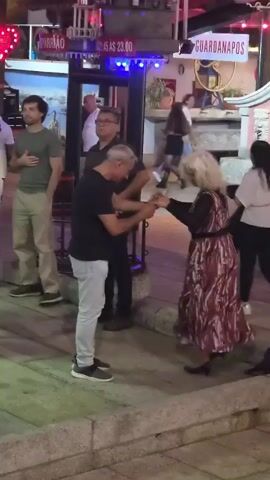 At 00:00, a man in a gray t-shirt and khaki pants stands to the left of the frame. Behind him, a glass-enclosed booth with blue lights is visible, with a sign reading "GUANABARA" and "AS 23.00". To the right of the booth, a woman with blonde hair in a patterned dress dances with a man in a black t-shirt and light gray pants. Another man in a black shirt stands behind them. In the background, a sign reads "GUARDANAPOS". At 00:01, the blonde woman raises her arms. At 00:02, she turns her back to the camera. At 00:03, she faces the camera again and continues dancing with the man. At 00:04, the couple continues to dance, with the man in the black t-shirt looking down.