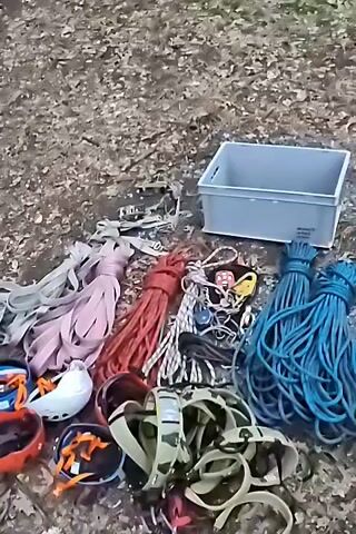 The video displays a collection of outdoor adventure gear spread out on a surface covered with fallen leaves and twigs, suggesting an outdoor setting like a forest floor. The items are arranged in a pile, with some items more clearly visible than others. Prominently featured are several ropes of varying colors, including pink, red, white, and blue. There are also multiple climbing harnesses, some with camouflage patterns, and several helmets, at least two of which are white and one orange. A grey plastic bin sits to the right of the gear. Scattered among the ropes and harnesses are various carabiners and other metal climbing accessories. The overall impression is of equipment prepared for activities such as rock climbing, rappelling, or tree climbing. The camera appears to be positioned above the scene, looking down at the collection of gear. There are no people or animals visible in the frame, and no actions or interactions are depicted.
