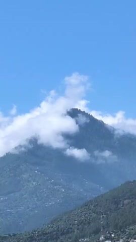 The video shows a view of mountains against a clear blue sky with scattered white clouds. The mountain peaks are covered in lush green vegetation. In the foreground, there is a town or village nestled on the slopes of the mountains, with buildings and houses visible. The overall scene depicts a scenic natural landscape with human settlement integrated into it.