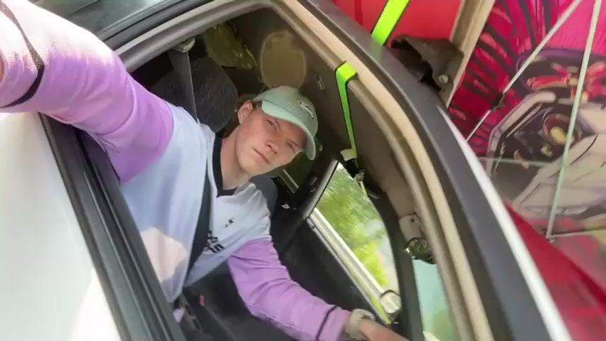 A young man wearing a light blue baseball cap and a white and purple long-sleeved shirt is seen through the open driver's side window of a white car. He is looking down and to his right. Outside the car, several red paddleboards are strapped to the roof rack. The paddleboards have pink and white graphic designs on them. The scene appears to be outdoors with greenery visible in the background.
