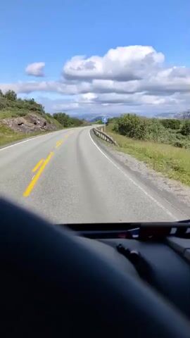 The video shows a view from inside a vehicle driving on a road in Sandnessjøen, Norway. The road is paved and has yellow lines marking the lanes. The vehicle is driving on the right side of the road, and there is a guardrail on the right side of the road, separating it from a body of water. In the distance, a large cable-stayed bridge can be seen, which the vehicle is approaching.

The scene is set during the daytime, with a bright blue sky and scattered white clouds. The weather appears to be good, with clear visibility. The surrounding landscape is mountainous and green, with trees and bushes dotting the hillsides. The water next to the road is calm and reflects the sky.

There are a few signs visible along the road. One sign indicates that a restroom is 800 meters ahead. Another sign shows a warning for curves. There is also a speed limit sign indicating 40 km/h.

No people are explicitly visible in the video, but there are other vehicles on the road, including a truck and a car. The overall impression is of a scenic drive through a beautiful natural landscape in Norway. The video captures the essence of a road trip, with the continuous movement of the vehicle and the changing scenery.