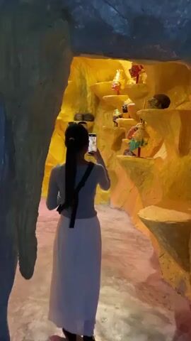 A person wearing a gray long-sleeved shirt and a white skirt stands with their back to the camera, holding a mobile phone and taking a picture of a display of masks. The masks are placed on tiered shelves carved into a rock-like structure that is painted yellow and orange. The masks appear to be of various colors and styles, some with ornate headdresses. The environment appears to be dimly lit, with a spotlight illuminating the masks. The floor is a light-colored concrete.