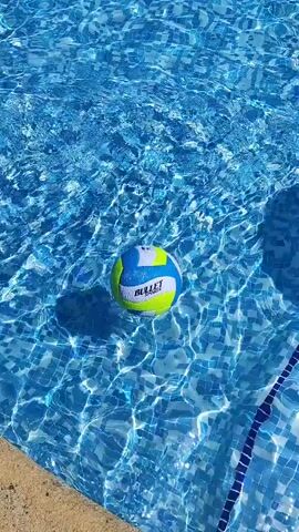 A blue, yellow, and white volleyball floats in clear blue swimming pool water. The ball is near the edge of the pool, which has a beige coping and a blue tiled line. Sunlight glints off the water's surface, creating ripples and reflections.