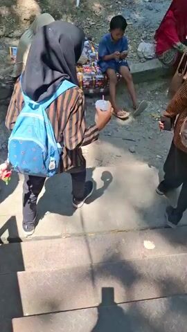 The video shows an outdoor gathering of people in a wooded area, likely a park. In the foreground, several individuals, mostly women wearing hijabs, are seated on the ground. One woman in a red and black striped shirt with a backpack is facing away from the camera, holding a cup with a straw. Behind them, a group of women in hijabs are sitting together, some engaged in conversation. Further back, more people are scattered throughout the park, with some children near a playground structure. There are trees providing shade, and the ground appears to be a mix of dirt and gravel. A small gazebo with an orange roof is visible in the middle ground. The overall atmosphere appears relaxed and casual, suggesting a social outing or event.
