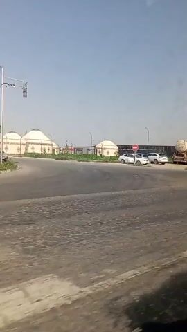 The video pans across a dusty road with several cars and yurts in the background under a clear blue sky. The yurts are round, white, and domed structures with decorative trim. Some of the yurts are clustered together, while others are more spread out. Several cars are parked in front of the yurts, including a silver sedan, a white sedan, and a silver SUV. There is also a large tanker truck parked on the right side of the frame. The road itself appears to be made of asphalt and is somewhat cracked and worn. There are some traffic lights and signs visible. The overall impression is one of a semi-arid environment with traditional nomadic dwellings.
