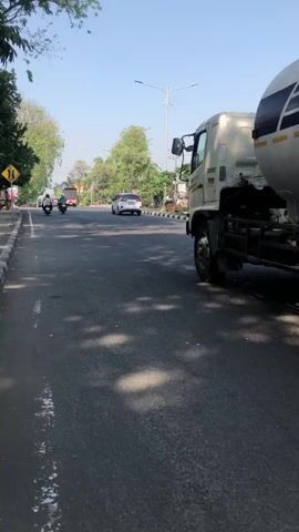 The video shows a street scene with a large tanker truck labeled "SAMATOR" driving on a paved road. A person on a motorcycle is riding alongside the truck, to its right. In the background, there are trees, power lines, and buildings. The sky is clear and blue. The overall impression is a busy road with various vehicles and infrastructure typical of a developed area.
