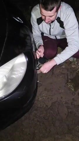 Two individuals are in a muddy, outdoor environment at night. One person is kneeling next to the front of a black car, appearing to attach a tow strap to it. Another person is standing nearby, and a third person is holding a tow strap. The car's headlight is illuminated. The overall scene suggests an attempt to tow or retrieve the car.