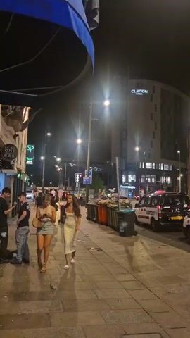 The video shows a city street at night. People are walking on the sidewalk, some in groups. The street is lit by streetlights and the lights from businesses. A taxi is driving by in the street. There is a building with the sign "Clayton Hotel" visible in the background. There are trash cans and other objects on the sidewalk. Two women are walking in the foreground, one looking at her phone. A blue awning is partly visible in the upper left corner of the frame.
