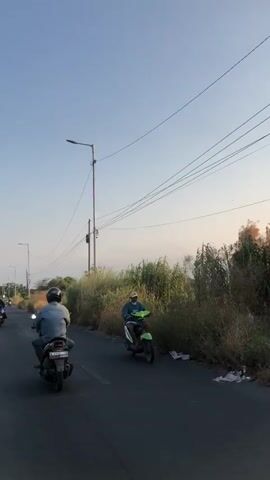 The video opens with a wide shot of a paved road bordered by dry vegetation and trees on the right. The sky is clear with a pale blue hue, suggesting it is either early morning or late afternoon. Overhead, utility wires stretch across the frame, supported by poles.

In the foreground, two individuals on motorcycles are traveling away from the camera on the right side of the road. The motorcycle closer to the camera is ridden by a person wearing a blue denim jacket and a dark helmet, facing away from the viewer. The motorcycle is dark-colored. Further ahead and to the right, another individual on a green motorcycle is also riding away from the camera. This rider is wearing a light-colored shirt and a light-colored mask, and a dark helmet.

The road appears to be empty of other vehicles at the moment, except for possibly another motorcycle partially visible in the far left background. The environment is rural or suburban, characterized by the roadside vegetation and the general atmosphere. The lighting indicates natural daylight.