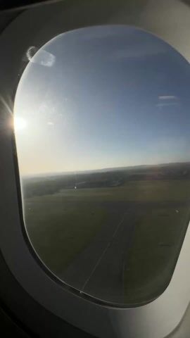The video shows a view from an airplane window. The sky is blue with a slightly hazy horizon. Below, there are green fields, patches of trees, and what appear to be buildings in the distance.  The plane seems to be flying fairly low, offering a clear view of the landscape.  There is some glare from the sun on the window.