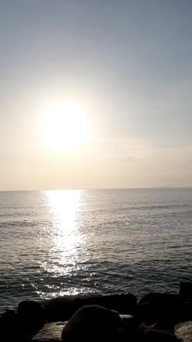 The video shows a view of the ocean at sunrise, with the sun reflecting on the water. There are rocks in the foreground and a small boat in the distance. The sky is mostly clear with a few clouds near the horizon.