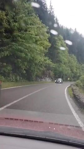 A car travels on a two-lane paved road through a mountainous region with lush green trees on both sides. The weather is overcast with raindrops on the windshield. The road curves ahead, and there's a small stone wall running along the right side. On the right, there is a river or stream that is full and appears to be carrying sediment. Another car approaches, driving in the opposite direction.