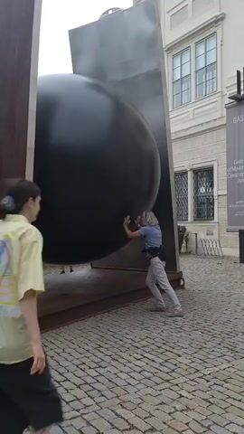 A person in a yellow shirt walks away from a large, black, spherical sculpture. Another person in a grey shirt and light-colored pants pushes against the sculpture. The sculpture is made of metal and is mounted on a stand. The ground is paved with cobblestones. A building is visible in the background.