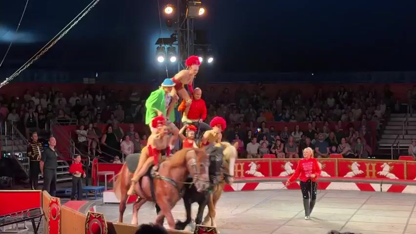 A circus performance is underway in a large tent with a packed audience. Three horses are lined up in a ring, each with performers on their backs. A pyramid of acrobats has been formed on the horses, with a woman in a red costume at the very top, balancing on one leg with her arms outstretched. Below her, a man in a red shirt is standing, and beneath him, a person in a green shirt and blue hat is seated. Several other performers are positioned on the horses, some in red costumes, one in a yellow and red costume, and one in a red and yellow costume. A ringmaster in a red jacket stands to the right, facing the performers. The audience is seated in tiered seating around the ring. The lighting is dim, with spotlights focused on the performers.