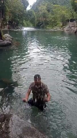 A man wades in a clear river surrounded by lush greenery. The water appears to be shallow enough that he can stand. He reaches out to a rock at the river's edge, pulls himself up and out of the water, and steps onto the shore. The area is a natural setting with trees and what looks like a small waterfall in the distance.