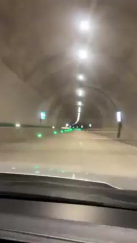 The video shows a vehicle driving through a tunnel. The tunnel has an arched ceiling with a row of lights running along its center.  Green lights are spaced intermittently along the walls.  The road is grey and has a single lane divider line.  The perspective is from inside the vehicle, looking forward through the windshield. The dashboard is visible at the bottom of the frame.  A musical track plays.