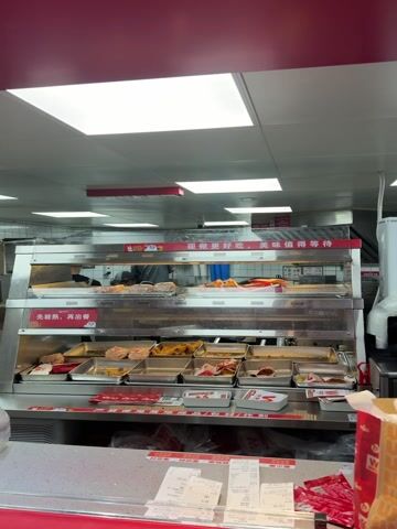 The video shows the interior of a fast food restaurant, likely a KFC. A food preparation area is visible behind a counter with a display case containing various food items like fried chicken, sandwiches, and other sides. A restaurant worker wearing a mask is visible in the background. Receipts and packaging materials are on the counter. A sign above the food display reads "现做更好吃，美味值得等待" (Freshly made is more delicious, the taste is worth the wait).