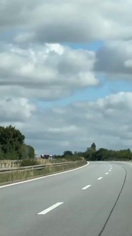The video shows a view from a moving vehicle on a highway. The highway has two lanes in the direction of travel, marked by white dashed lines. On the left side of the highway, there is a metal guardrail. Beyond the guardrail, there is vegetation, including trees and tall grass. In the distance, several trucks are parked on the shoulder or a nearby area off the highway. The trucks are of various colors, including white, black, and orange. The sky is partially cloudy with patches of blue sky visible. The overall impression is a typical highway scene on a cloudy day.