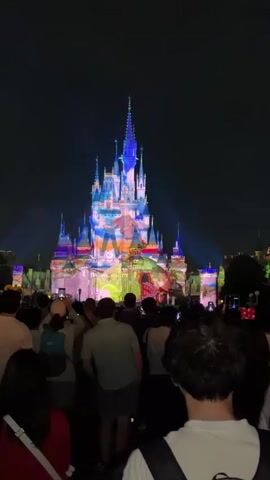 The video captures a nighttime scene in front of a castle. A large crowd of people is gathered, facing the castle, with many holding up phones to record or take pictures. The castle itself is illuminated with a vibrant projection show, featuring various colors and patterns. Fireworks are being set off in the sky above the castle, with trails of light visible against the dark night sky. The overall atmosphere appears to be festive and celebratory, likely at a theme park or a similar event.