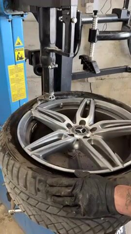 A gloved person is shown working a tire off of a Mercedes rim using a tire machine. The machine has a blue base, a black arm, and a metal piece that separates the tire from the wheel. The wheel has a silver finish and a prominent Mercedes-Benz logo in the center. The tire itself is black and appears used, with a visible tread pattern. The environment appears to be a workshop or garage, with various tools and equipment visible in the background. The person uses the machine to push the tire away from the wheel.