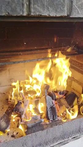 The video shows a close-up view of a fire burning in a barbecue grill. Logs and embers are visible, with bright orange and yellow flames engulfing them. Sparks are seen rising from the fire. The grill appears to be made of stone or brick, with a metal rack positioned above the flames. The overall scene is dimly lit, with the fire being the primary source of light.