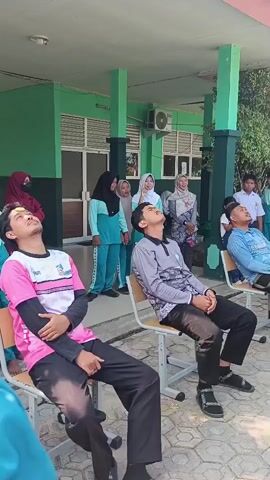 The video shows a group of young people, mostly teenagers, engaged in an outdoor activity. Several individuals, predominantly male, are seated on chairs with their heads tilted back, looking upwards. One young man in a pink and black shirt and black pants appears to be the focus of attention, with others looking at him. A young woman in a teal outfit and hijab is standing behind him, and another person in a light blue outfit is also looking up. In the background, other students and adults are gathered. Some are standing and observing, while others are interacting. The environment appears to be an open area outside a building with green walls and pillars. The ground is paved with interlocking bricks. The overall atmosphere suggests a game or a group activity, possibly a school event. The timestamp at the beginning indicates the beginning of the recording.