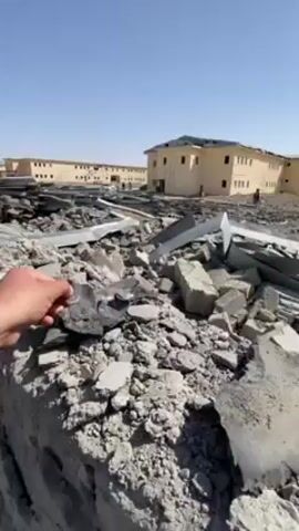 The video shows a man holding a piece of dark, rough material, standing in a debris-filled area. The ground is covered with scattered concrete blocks, metal sheets, and other wreckage. In the background, there are several low, light-colored buildings, some with visible damage like missing windows and holes in the walls. A few people can be seen moving around near the buildings. The sky is clear and blue. A white vehicle is parked in front of one of the buildings. The overall scene suggests the aftermath of an explosion or other destructive event.
