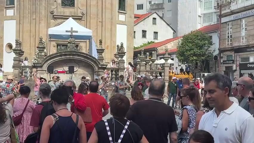 The video shows a crowd of people gathered in an outdoor public space, likely a plaza, in front of a large stone building with architectural detailing and a prominent cross on top, possibly a church or a historical monument. The crowd is diverse, with men, women, and children of various ages. Many are dressed casually in summer clothing. In the background, a group of people dressed in colorful shirts, some holding musical instruments, are visible. There is a white tent with a blue tarp behind them, and some audio equipment such as speakers and a microphone stand are present. The people in the crowd are facing the musicians, and some appear to be observing or participating in an event. The environment suggests a sunny day, with buildings and architecture typical of a European city. Some individuals in the foreground are looking towards the camera, while others are engaged with the activity happening in the plaza. The scene appears lively and festive.